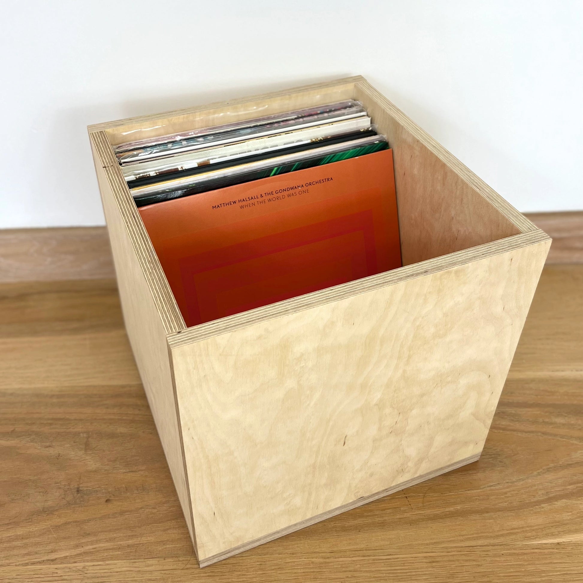 Square shaped wooden crate with pale thick side, sits facing diagonally to the right on a wooden floor. Records are displayed inside.