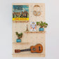 A large pale wooden pegboard with many drilled holes, four shelves & pegs faces front on. Various turquoise accessories and a ukulele are displayed on the board.