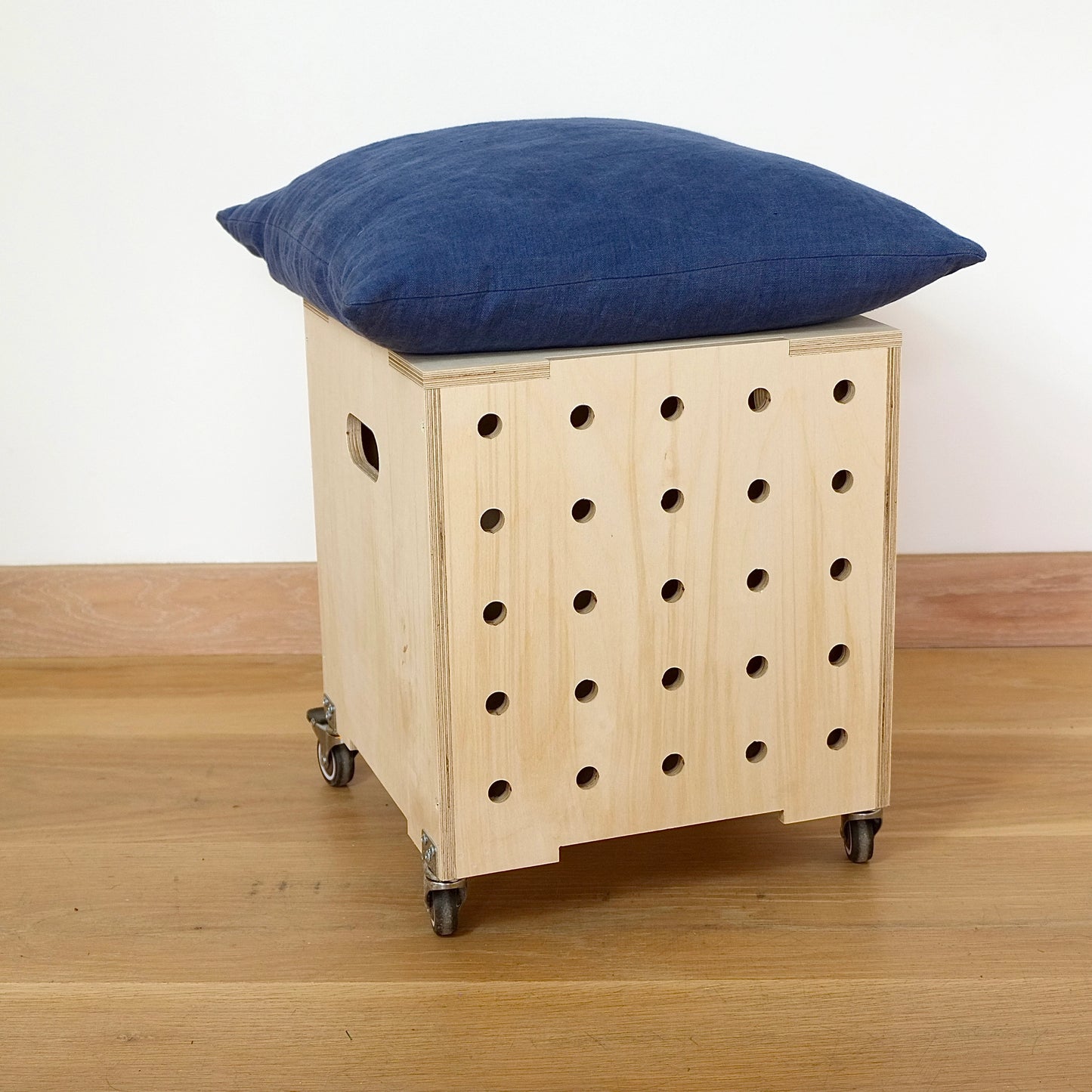 A simple pale wooden square shaped storage crate, faces diagonally to the right and sits on a wooden floor. There are five rows of holes cut in the front facet, it has castors and a lid with cushion on top.