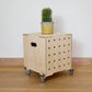 Simple pale wooden square shaped storage crate facing diagonally to right, with five rows of holes cut in front facet, castors, a wooden lid with cactus on top and sits on a wooden floor.