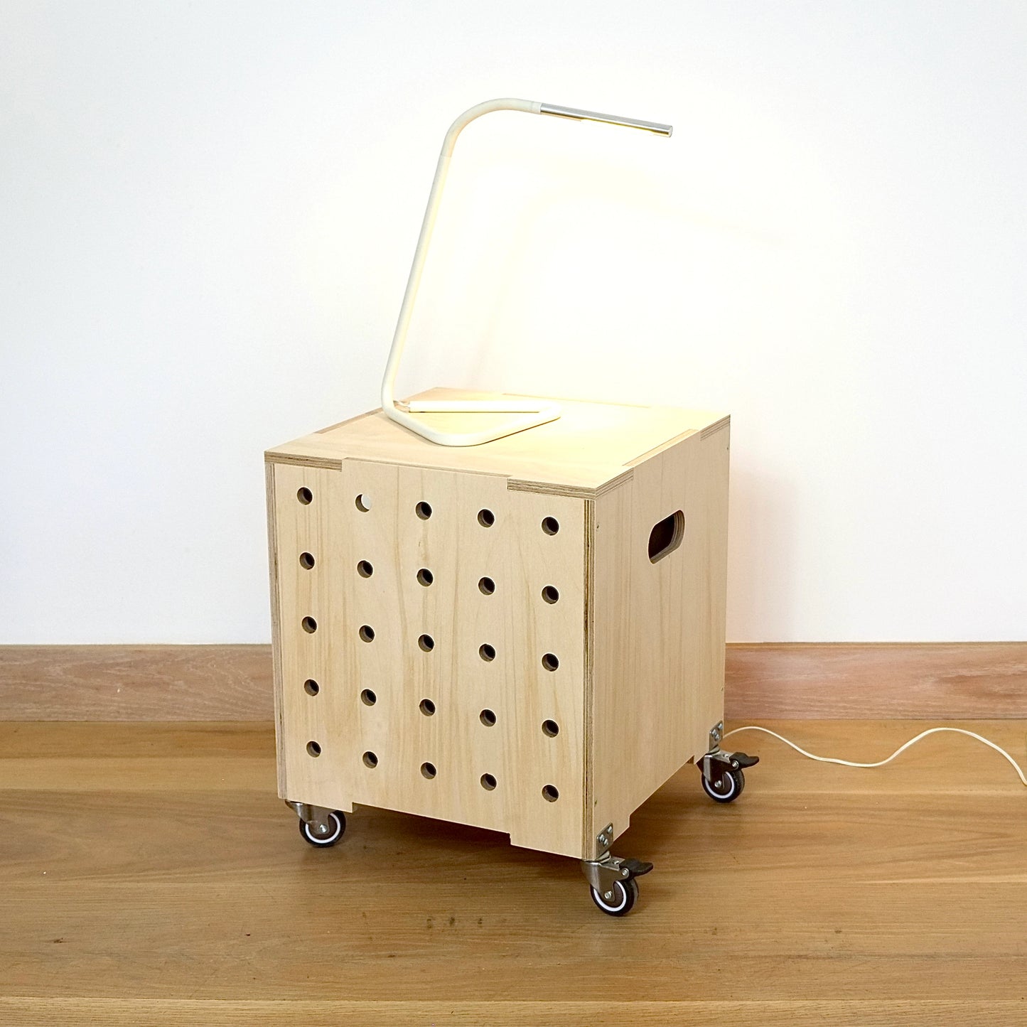 Contemporary birch plywood square box used as a side table with white lamp. front has 5 rows of vertical drilled holes, top has lid & bottom features castors against a white wall on a wooden floor