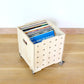 A Birch plywood open record crate containing vinyl records with 5 rows of drilled holes running vertical down front & castors on the base, standing on a wooden floor against a white background.