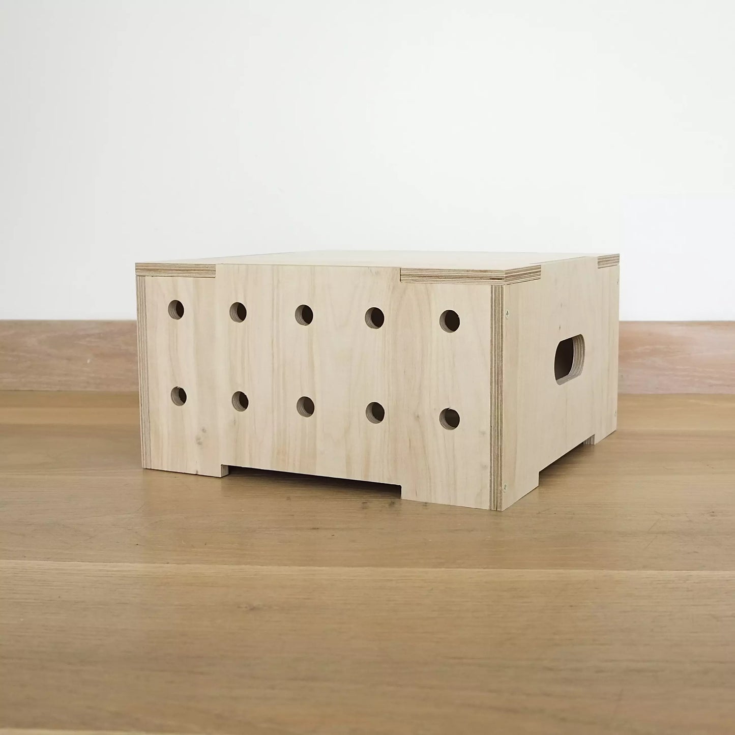 A modern pale wooden square crate with two rows of holes in front, with lid. It faces diagonally to left and sits on wooden floor.