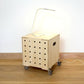 Simple pale wooden square shaped storage crate facing diagonally to left, with five rows of holes cut in front facet, castors, a wooden lid with lamp on top and sits on a wooden floor.