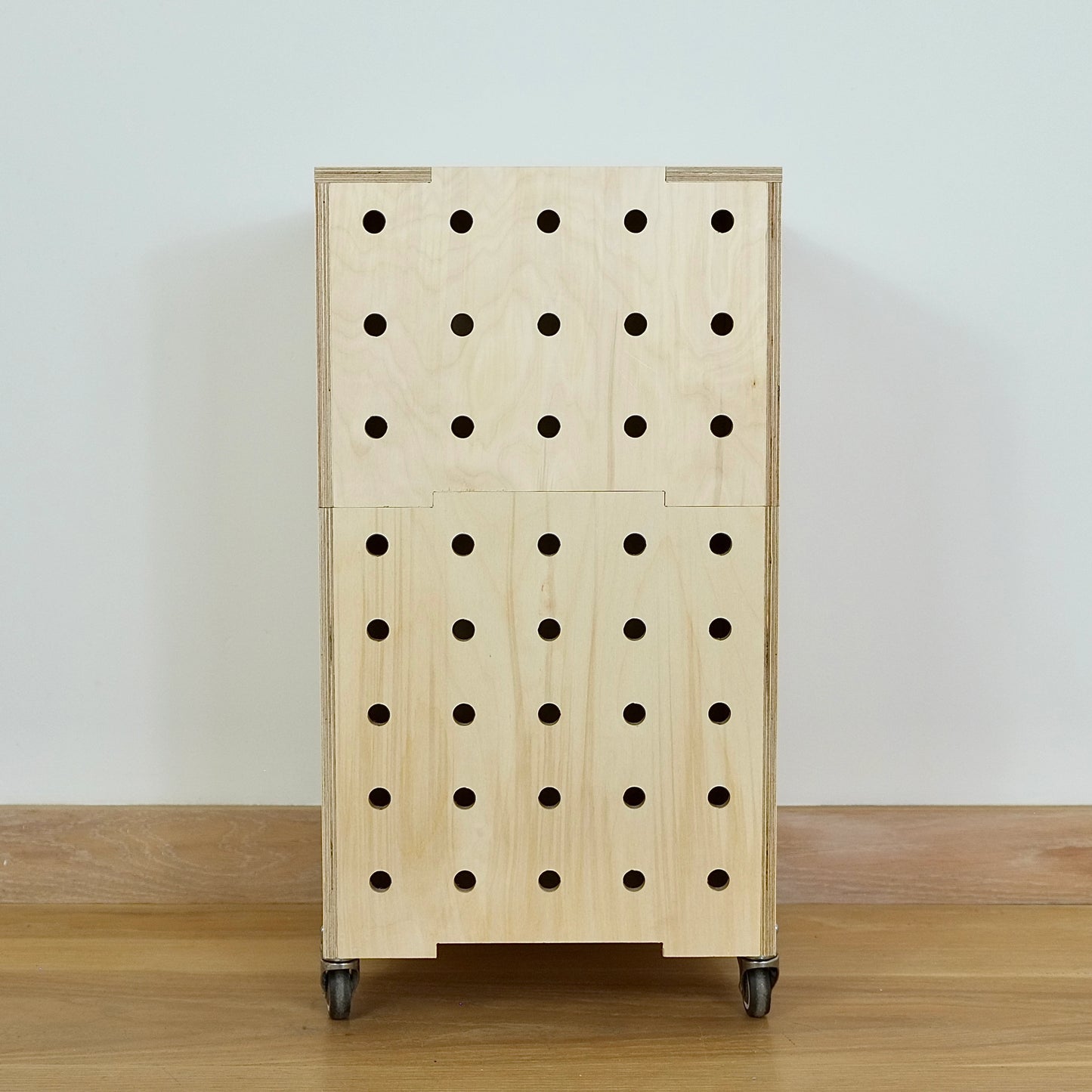 Modern plywood set of two stacking crates with 5 rows of vertical holes, castors & lid sitting on a wood floor against a white wall.