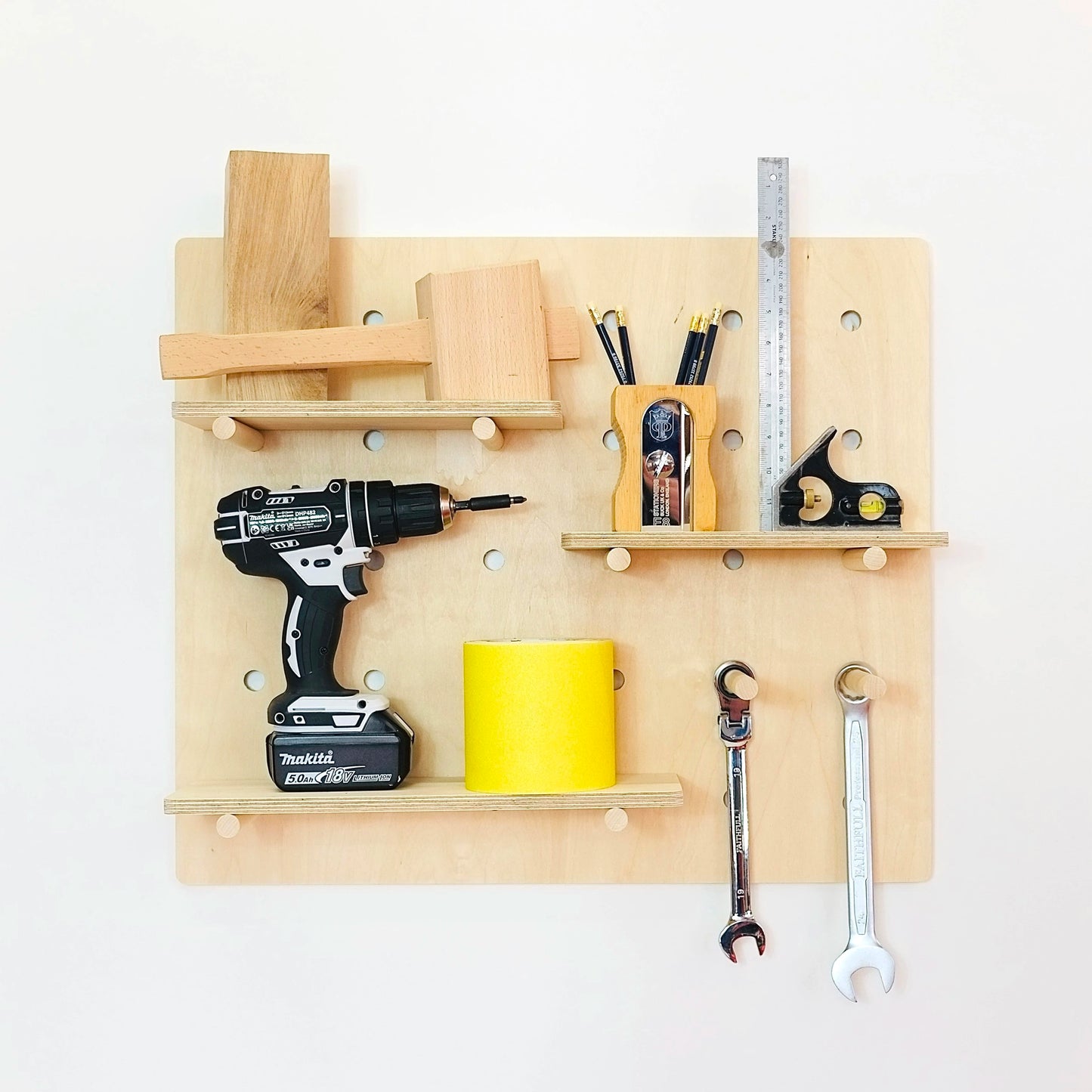 A medium pale wooden pegboard with many drilled holes, three shelves & pegs faces front on. Various woodworking tools are displayed on the board.