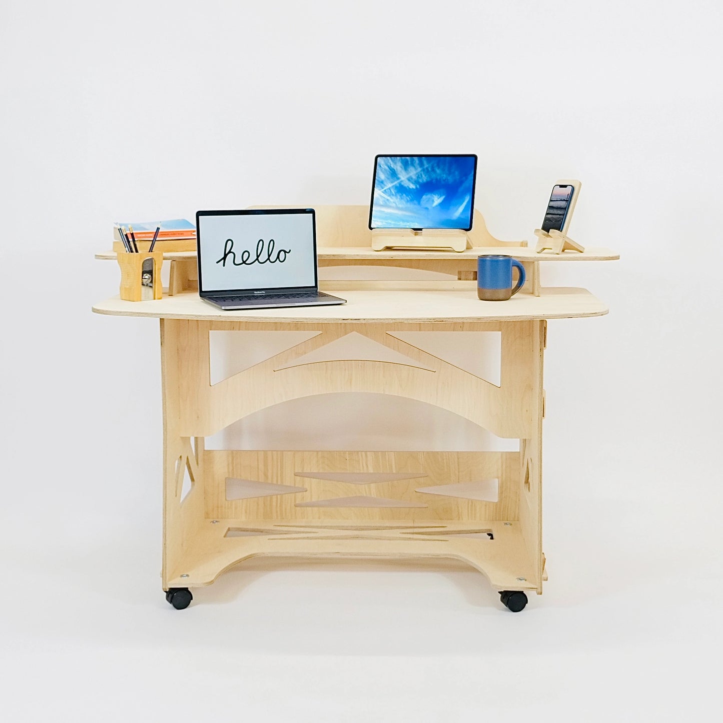 A pale wooden desk sits facing front on, it has castors. It has two computers, a mug, a pen pot , books and phone on stand sitting on top of it. 