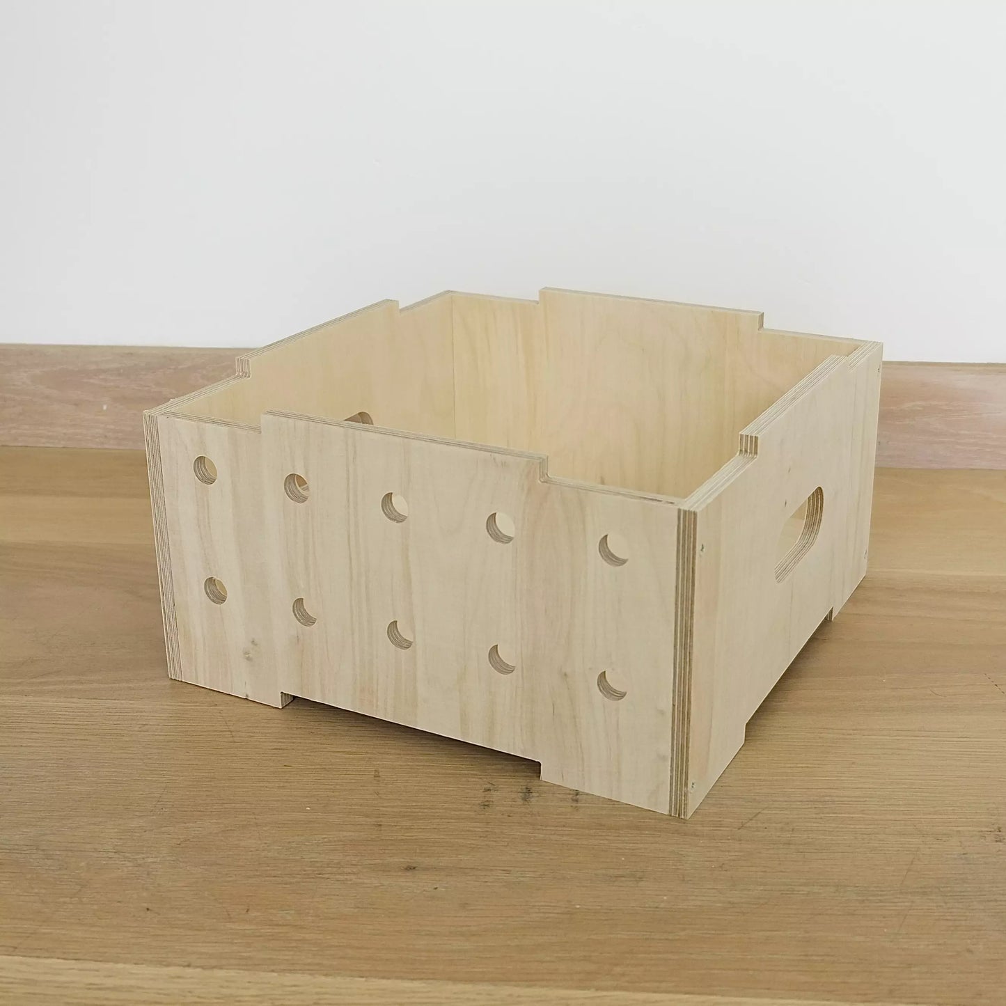 A simple pale wooden crate with two rows of holes in front. It faces diagonally to left and sits on wooden floor.