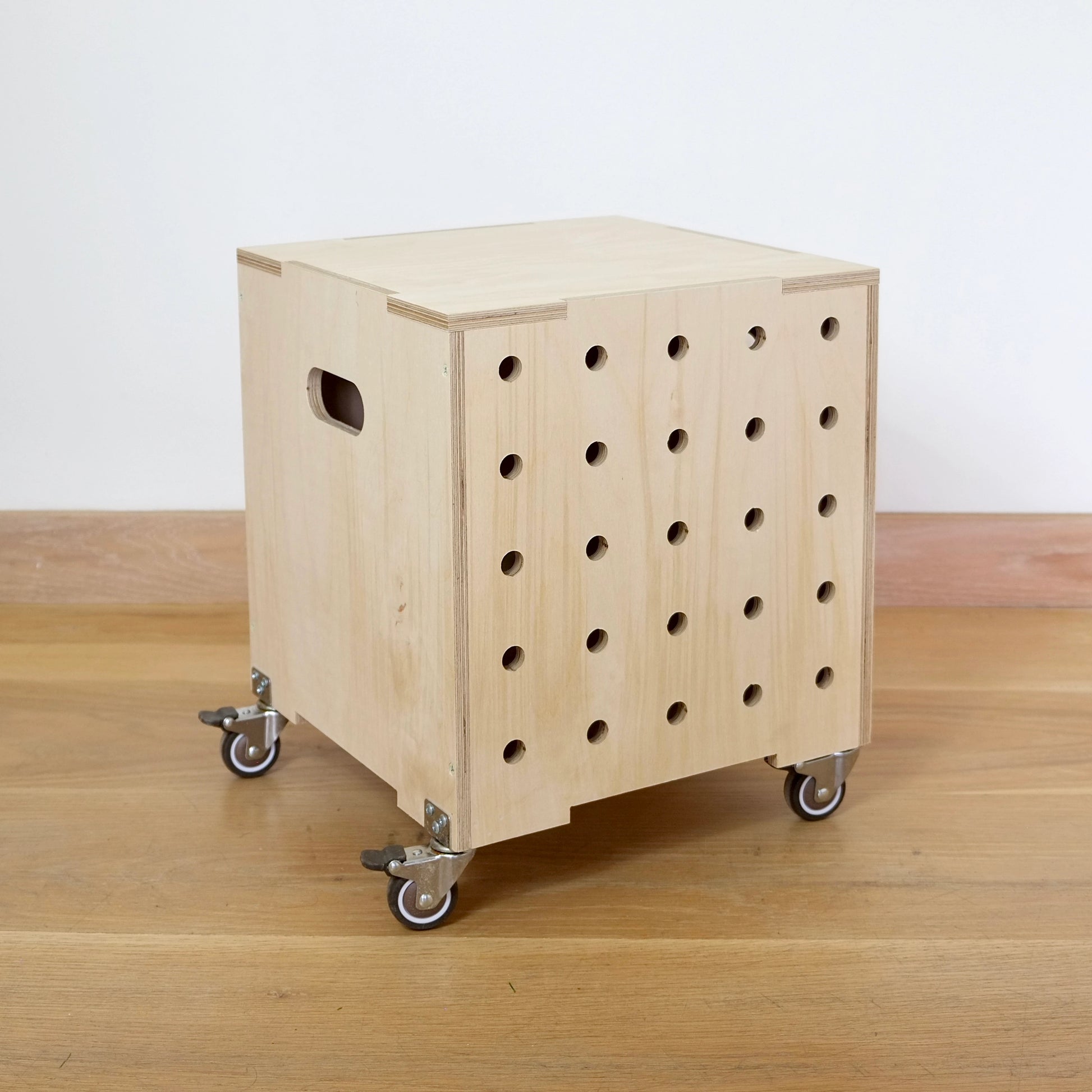 A simple birch plywood square shaped storage crate, faces diagonally to the right and sits on a wooden floor. There are five rows of holes cut in the front facet, it has castors and a lid.