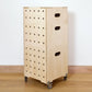 A stack of three simple pale wooden square shaped storage crates faces diagonally to the left, with ten rows of holes cut in the front facet, on castors, with a wooden lid and sits on a wooden floor.