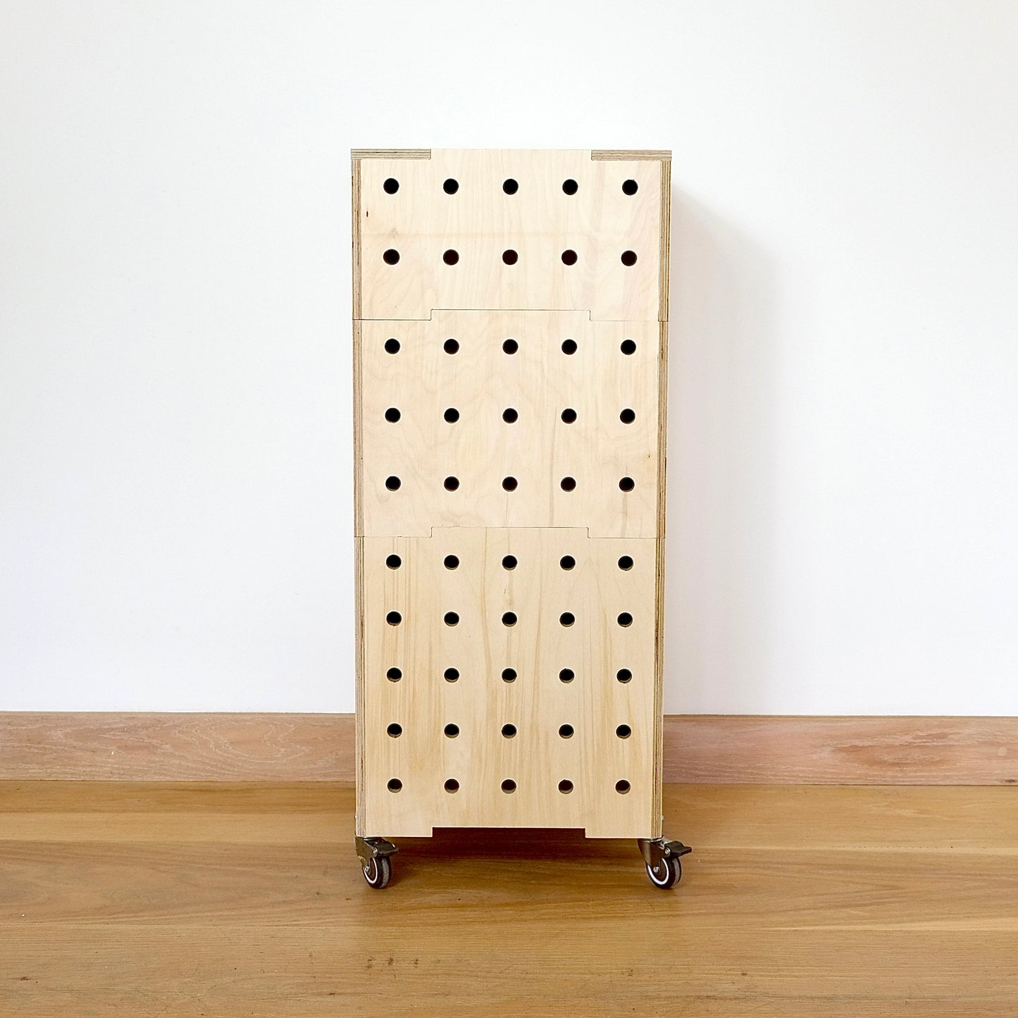 Birch plywood set of 3 stacking crates faces forward with 5 rows of vertical drill holes, 4 castors on base & wooden lid standing on a wood floor against a white wall.