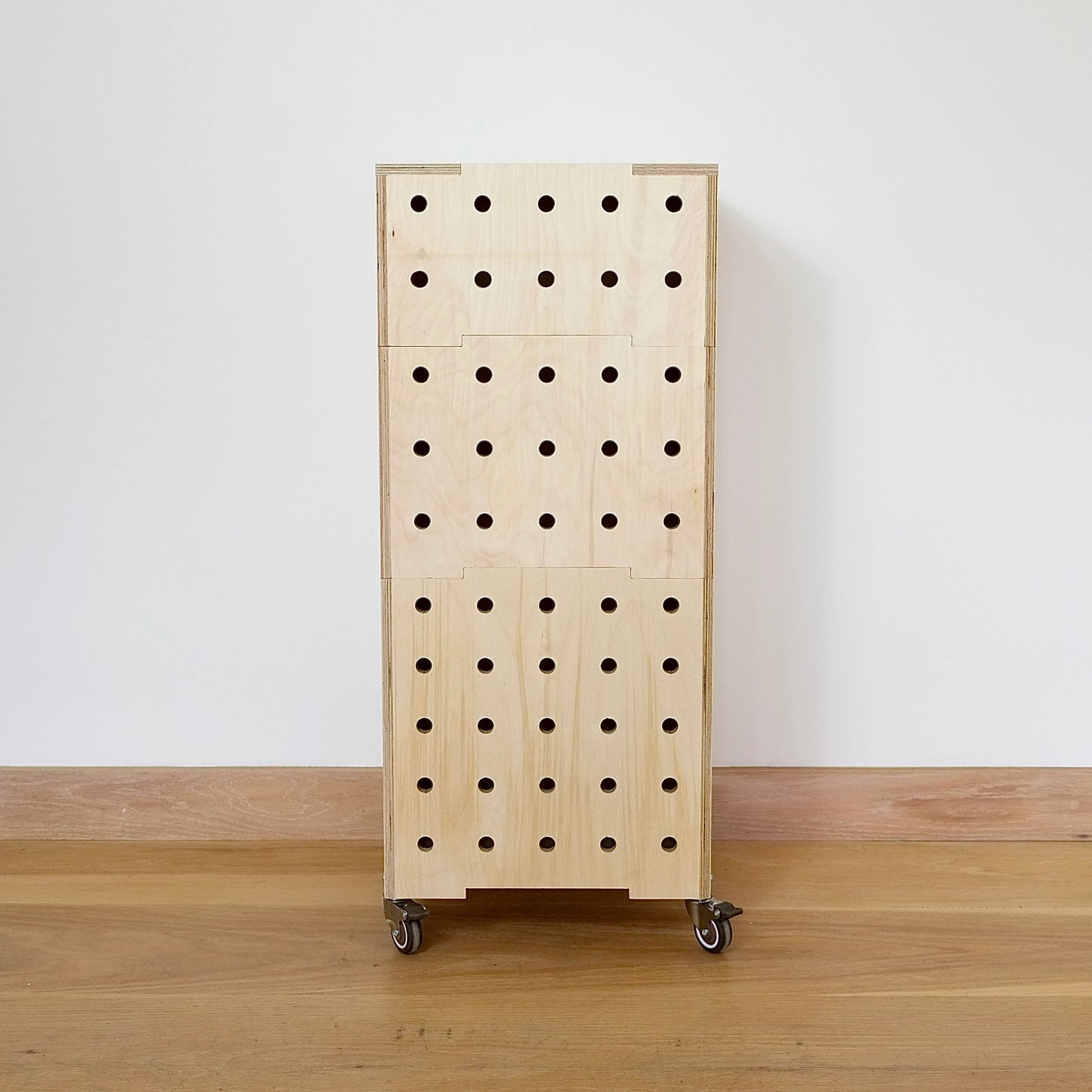 A set of 3 stacking pale birch plywood crates with 5 rows of drilled holes running vertical. The crates are set on 4 castors on a wooden floor against a white wall