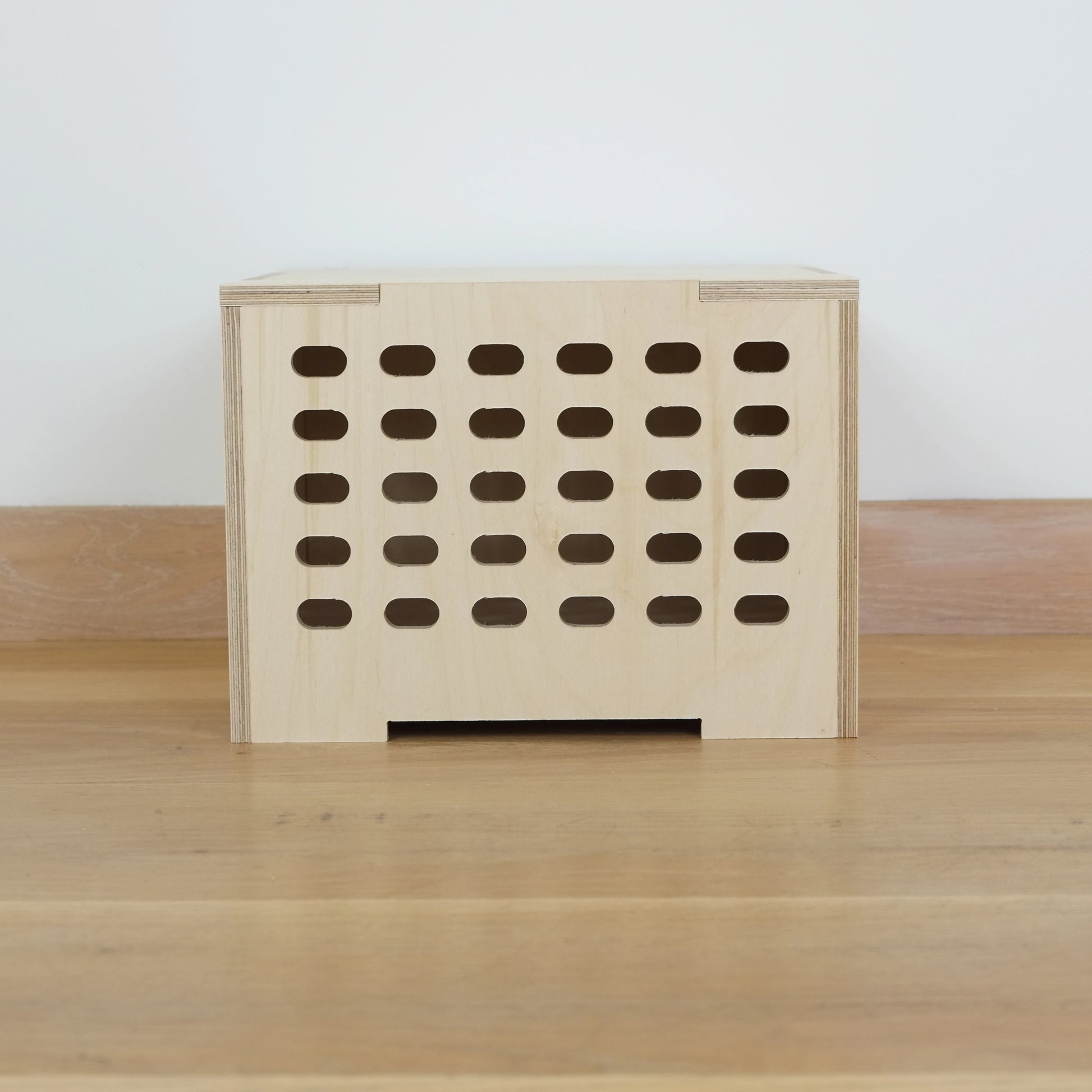 Birch plywood wooden crate with 5 rows of lozenge shape cutouts running vertically & wooden lid against a white wall