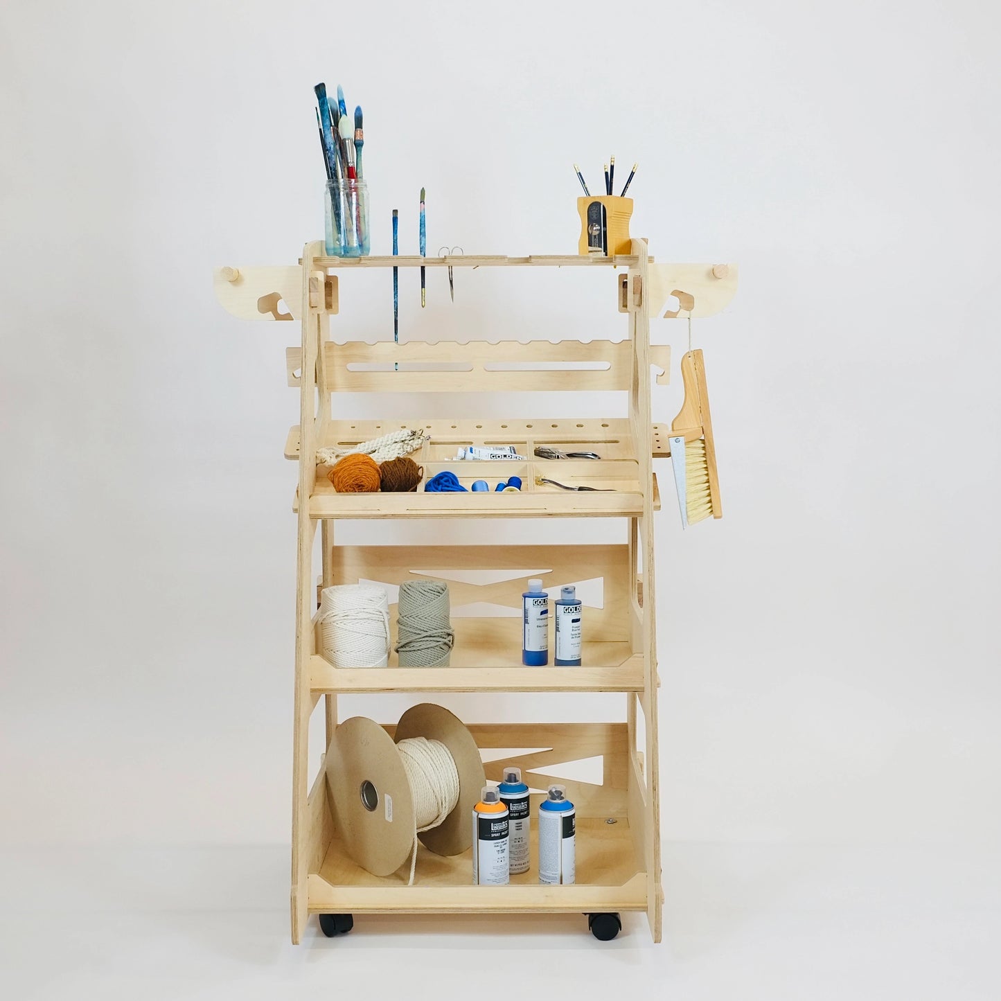 Pale birch plywood tool trolley with 3 shelving trays containing art supplies , rope & wool in tray dividers. Dust pan & brush hanging off wooden side hooks. Tool trolley on 4 castors facing forward against a white wall.