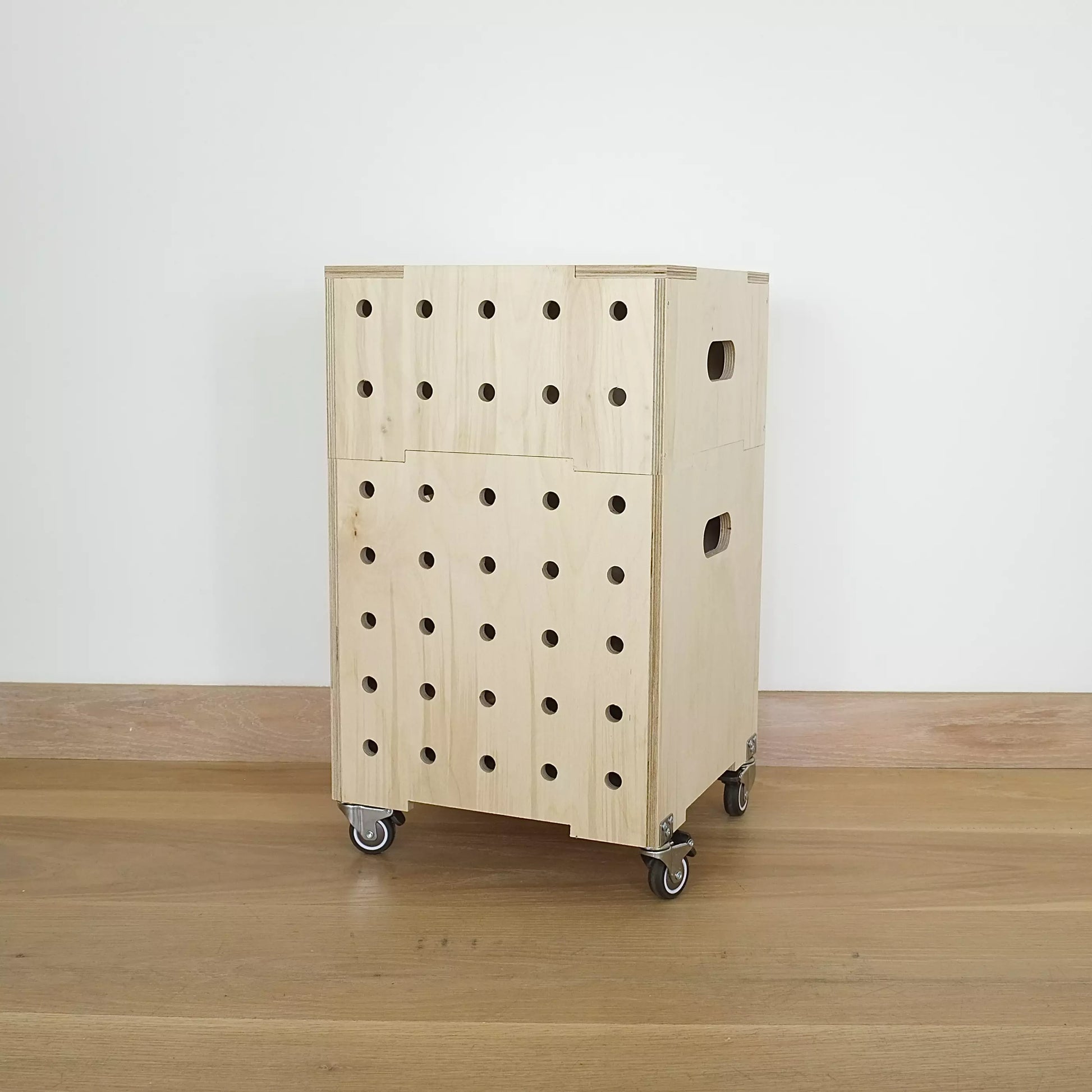 A stack of two simple pale wooden square shaped storage crates faces diagonally to the left, with seven rows of holes cut in front facet, castors and a wooden lid, sitting on a wooden floor.