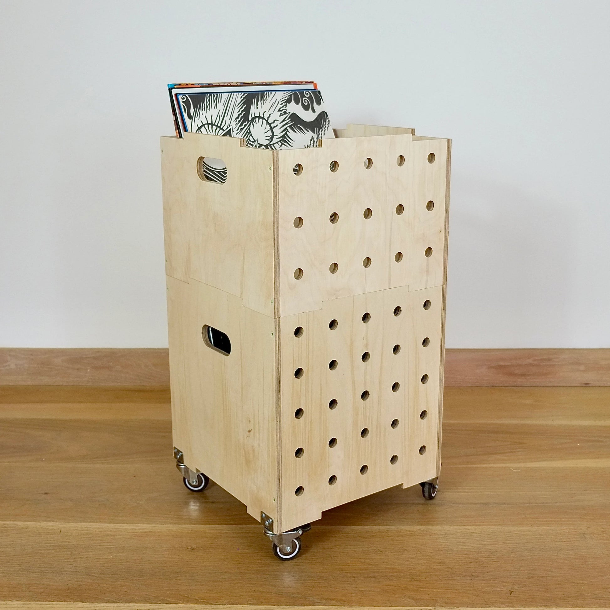 A stack of two modern plywood square shaped storage crates, faces diagonally to the right and sits on a wooden floor. There are eight rows of holes cut in the front facet, it has castors and displays records in the top crate.