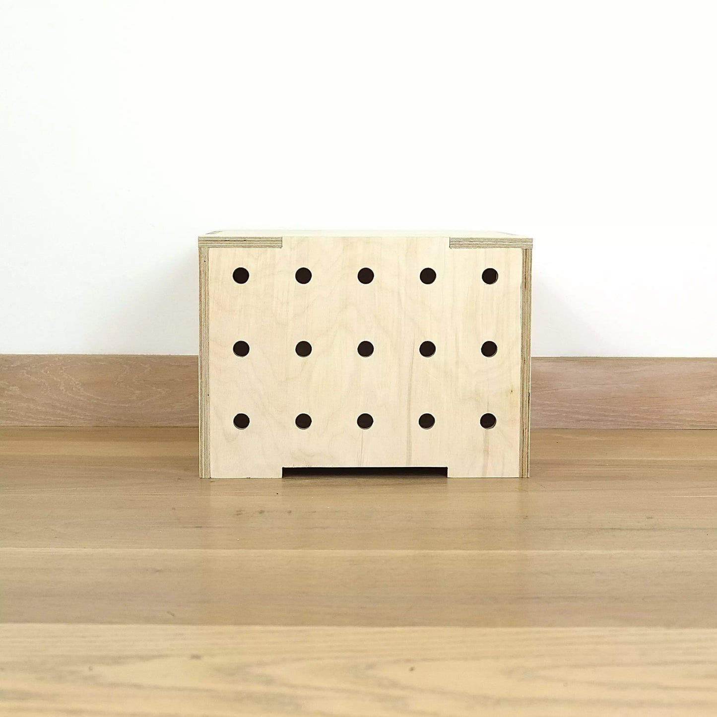 A simple pale wooden square shaped storage crate facing front on, with three rows of holes cut in the front facet and a wooden lid, sitting on a wooden floor.