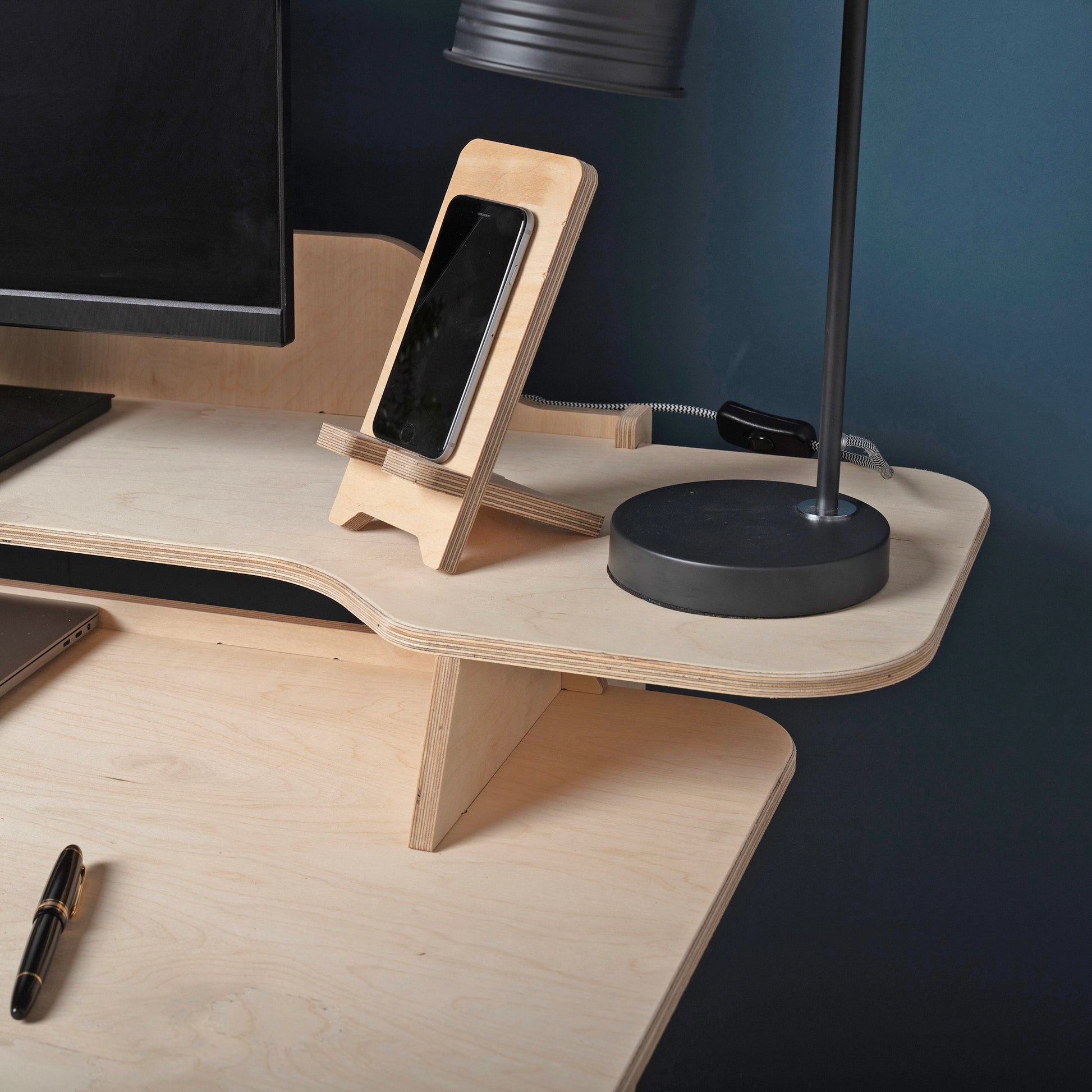 A close up showing details of pale wooden desk, including layers of plywood, and two desk top levels. A lamp, computer and phone stand sit on top of table. 