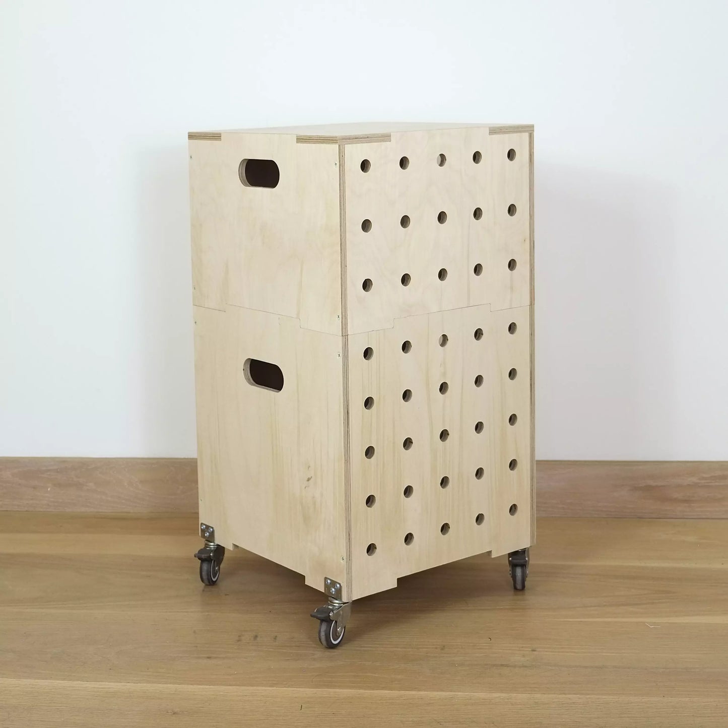 Stack of two simple pale wooden square shaped storage crates facing diagonally to right, with eight rows of holes cut in front facet, castors and a wooden lid, sitting on a wooden floor.
