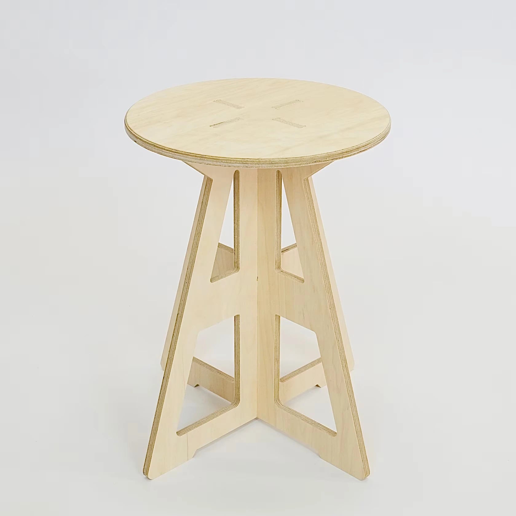 A simple modern pale wooden small stool. It has a round top and triangle legs which are slotted together in a cross shape, the base is wider than the top. The top also features four grooves where the legs slot into the top.