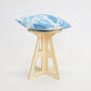 A simple modern pale wooden small stool. It has a round top and triangle legs which are slotted together in a cross shape, the base is wider than the top. The top also features four grooves where the legs slot into the top. A blue cushion sit on top.