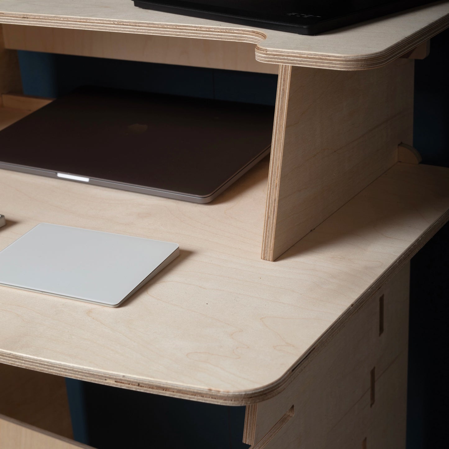 Close up for a two tiered birch plywood stand up desk with a laptop & note pad on desktop against a dark wall