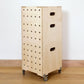 A modern set of pale birch plywood square box crates set at an angle against a white wall. The crates have 5 rolls of drilled holes running vertically down the front, hand holes on the sides & wheels at the base.