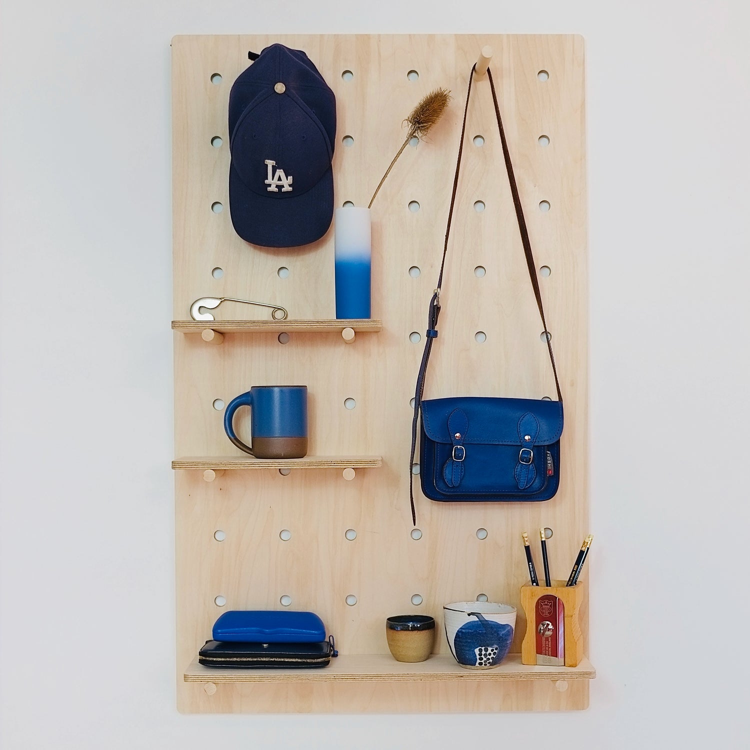 A large pale wooden pegboard with many drilled holes, three shelves & pegs face front on. Various blue accessories are displayed on the board.
