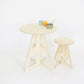 A simple modern pale wooden small table top and legs. It has a round top  and triangle legs which have slotted together in a cross shape with a matching stool. An orange, a small mug and bowl sit on the table.