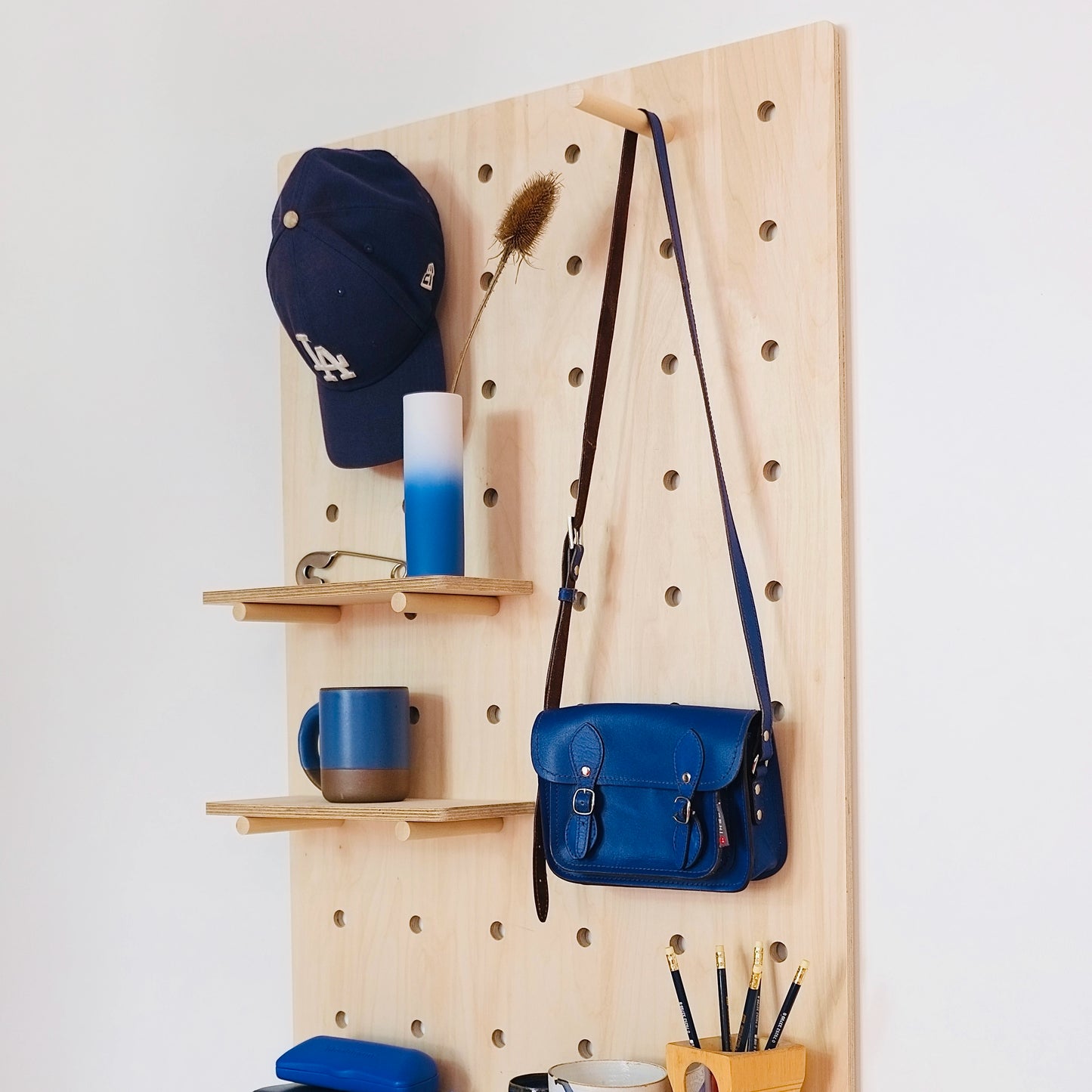 A large pale wooden pegboard with many drilled holes, two shelves & pegs faces diagonally to the left. Various blue accessories are displayed on the board.