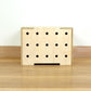A simple pale wooden square shaped storage crate facing front on, with three rows of holes cut in the front facet and a wooden lid, sitting on a wooden floor