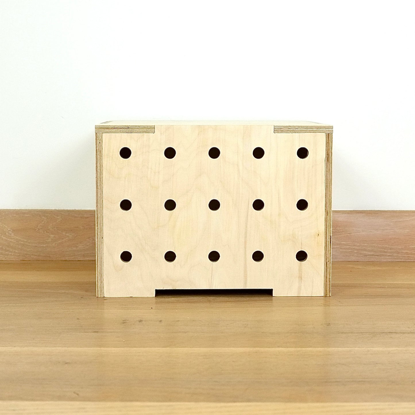 A simple pale wooden square shaped storage crate facing front on, with three rows of holes cut in the front facet and a wooden lid, sitting on a wooden floor