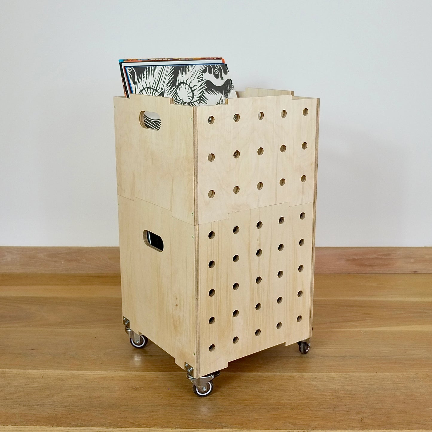 Open topped set of 2 pale wood birch ply stackable square crates with 5 vertical rows of holes on one side & hand holes on the other. It sits on 4 castors & holds a selection of vinyl records whilst standing on a wood floor with a white wall behind