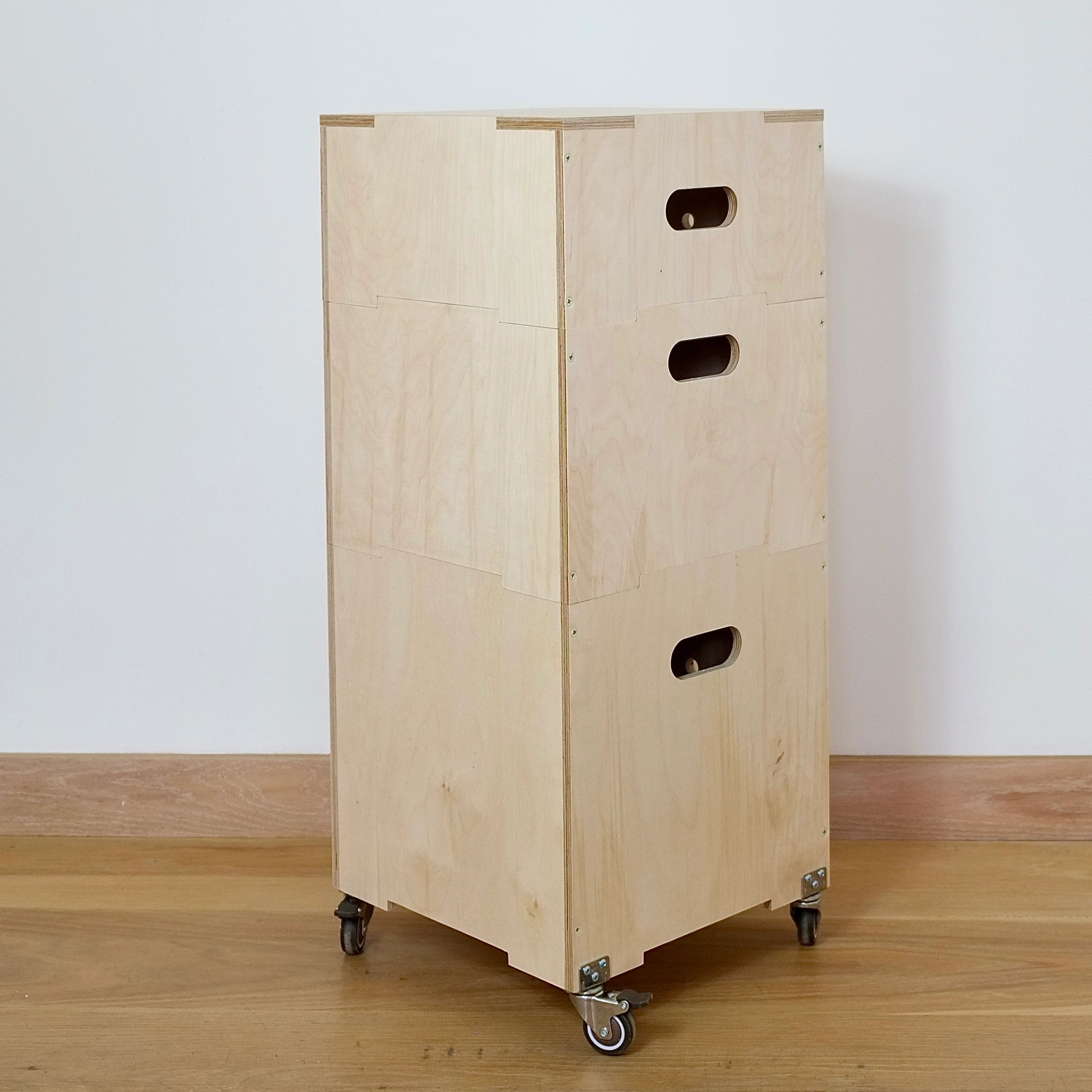 Set of three modern pale wood stackable square crates with hand holes, wooden lid & castors on base sitting on a wooden floor