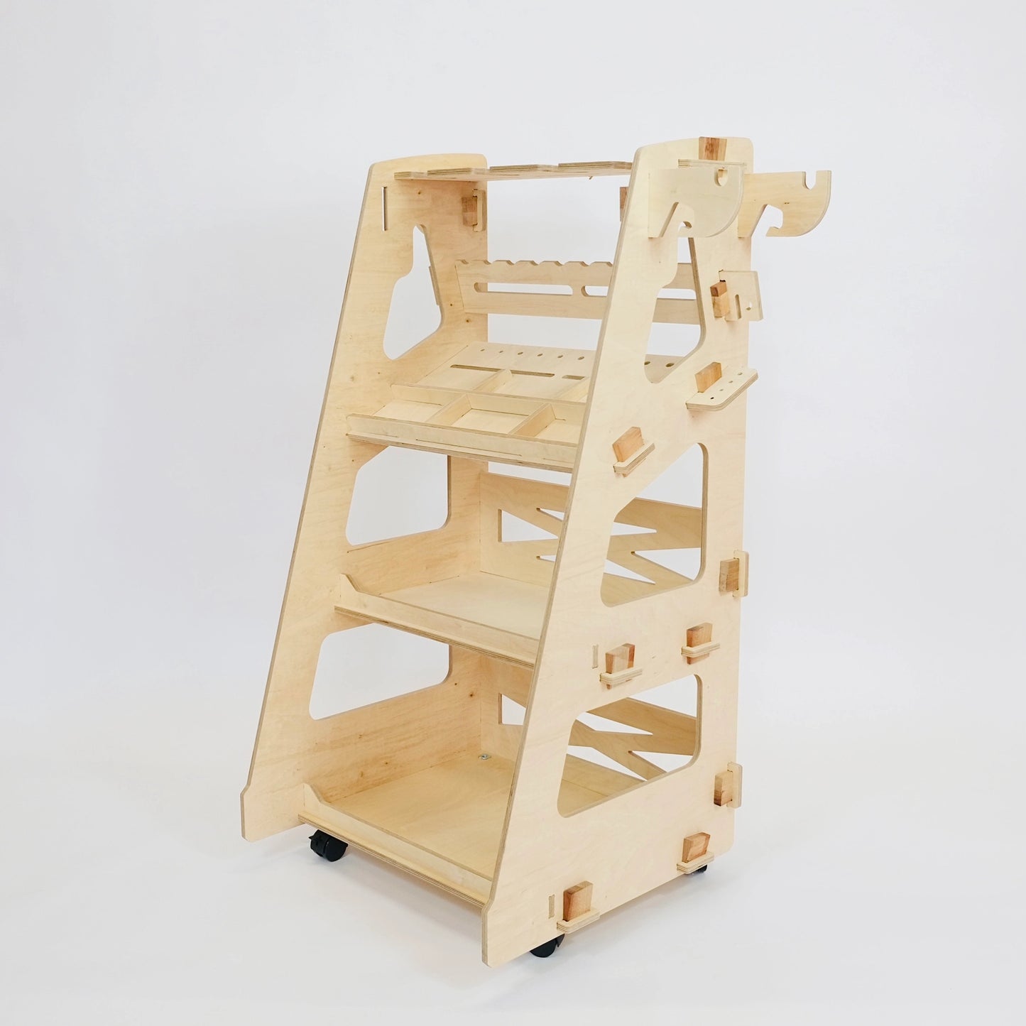 Pale birch plywood tool trolley with 3 slanted shelves, tool storage slots, 4 castors, cut out wood detailing on sides at an angle facing left on a white background.