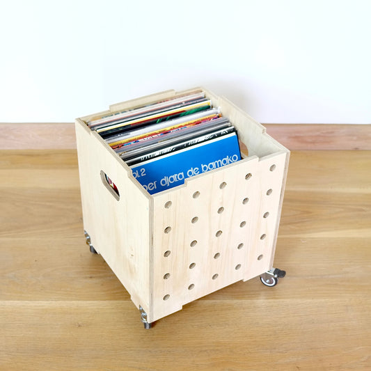 A simple pale wooden square shaped storage crate, faces diagonally to the right and sits on a wooden floor. There are five rows of holes cut in the front facet, it has castors and displays records in it.