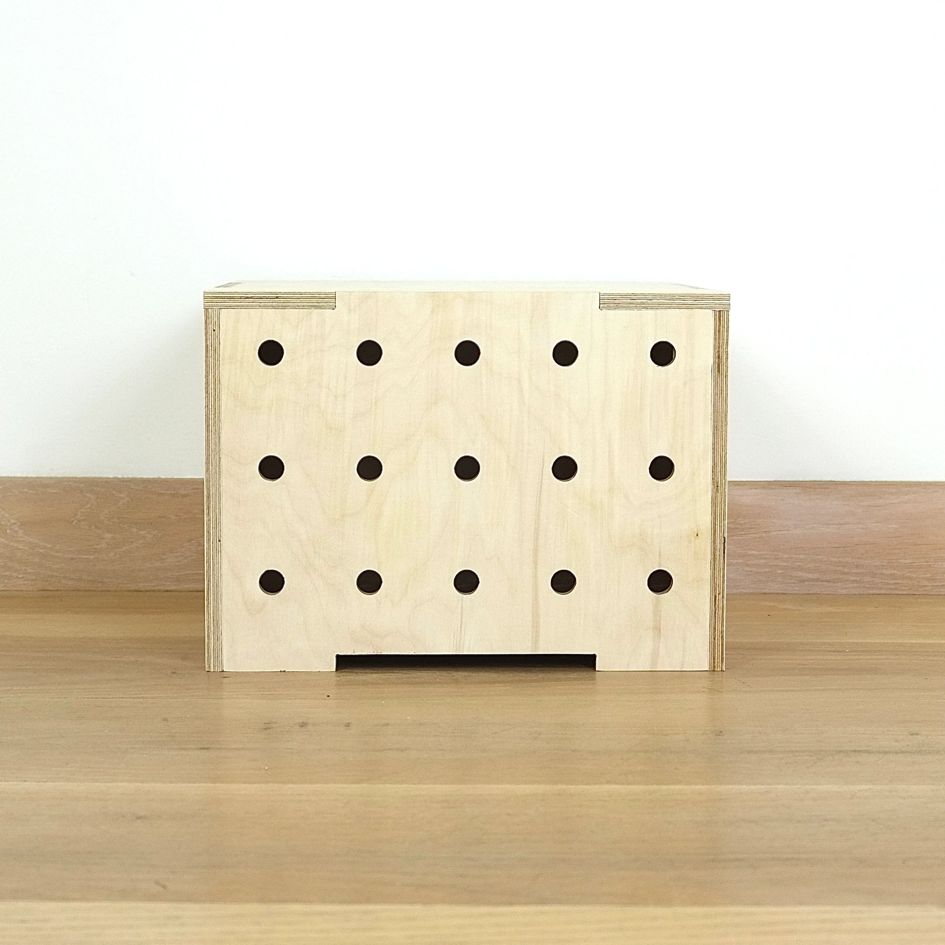 A simple pale wooden square shaped storage crate facing front on, with three rows of holes cut in the front facet and a wooden lid, sitting on a wooden floor.
