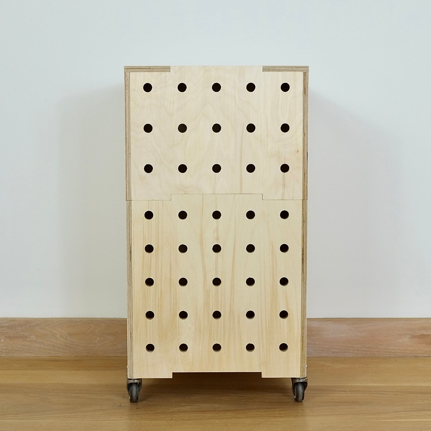 Stack of two simple pale wooden square shaped storage crates facing front on , with eight rows of holes cut in the front facet, castors and a wooden lid, sitting on a wooden floor.