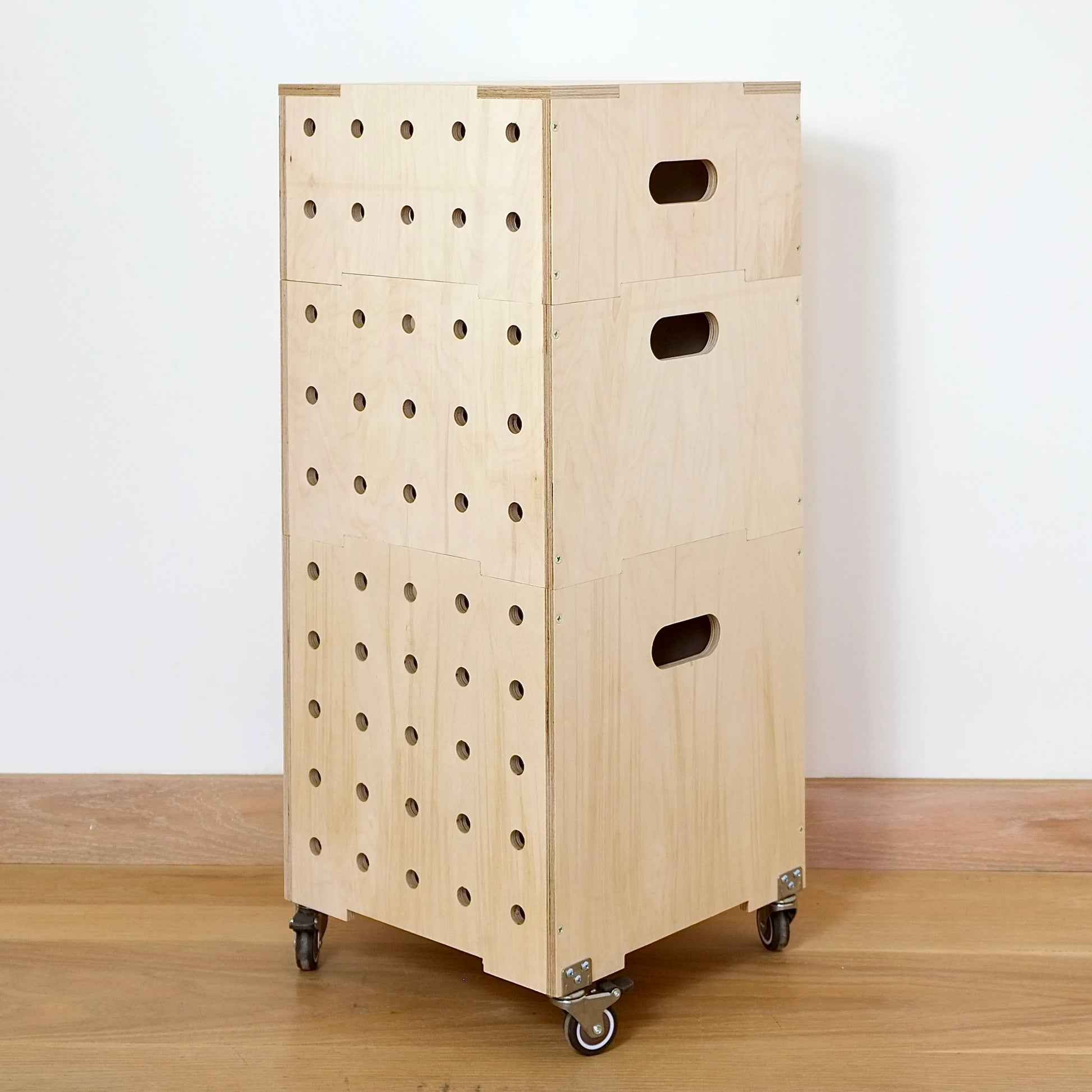 A tall stack of three pale plywood square crates with five rows of vertical holes on one side & hand holes on the other. It stands on four castors on a wooden floor infront of a white wall.