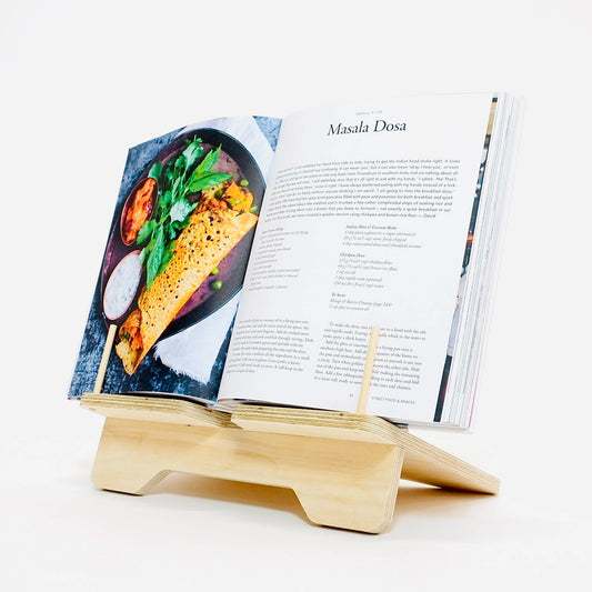 A pale wooden book stand faces diagonally to the left. An opened cook book sits open on the stand the pages are held back by two small upright dowels.