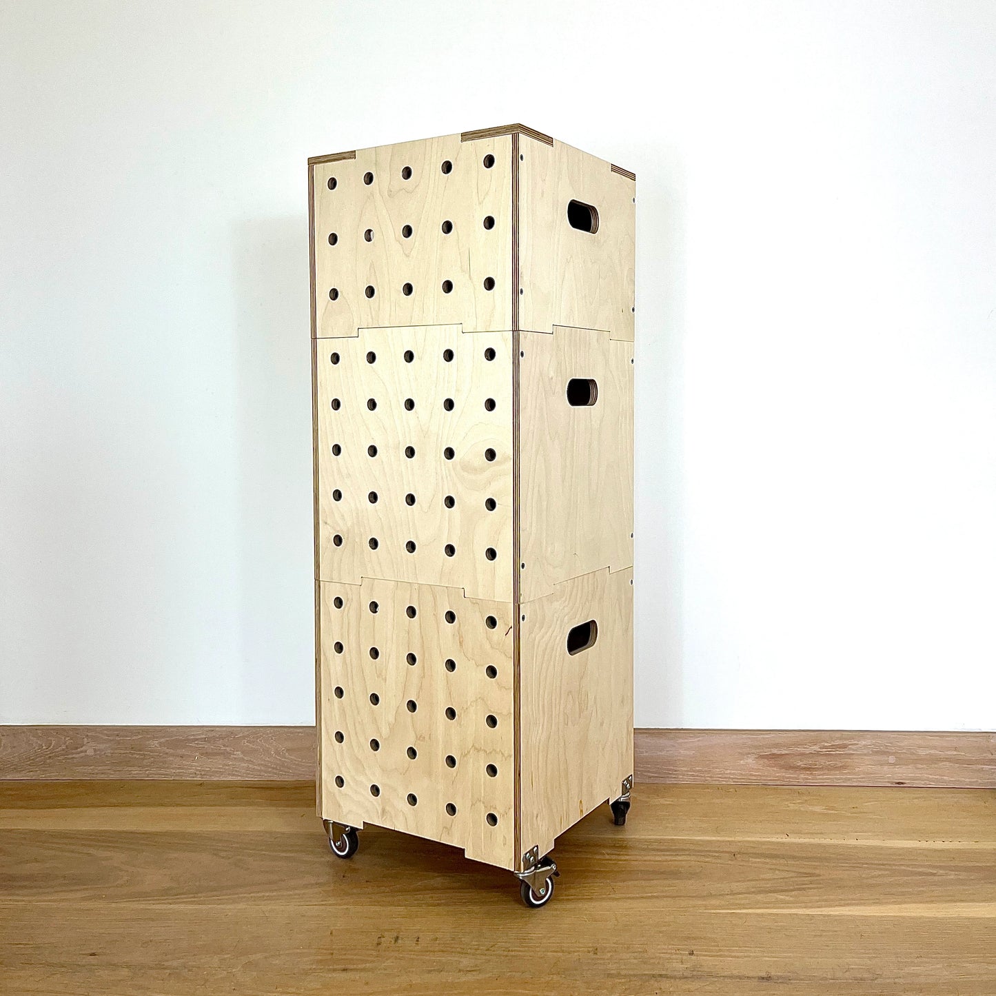 Stacking set of 3 birch wood ply square vinyl storage crates with 5 rows of vertical drilled holes on one side, hand holes on the other sitting on castors on a wood floor against a white wall facing to the left at angle