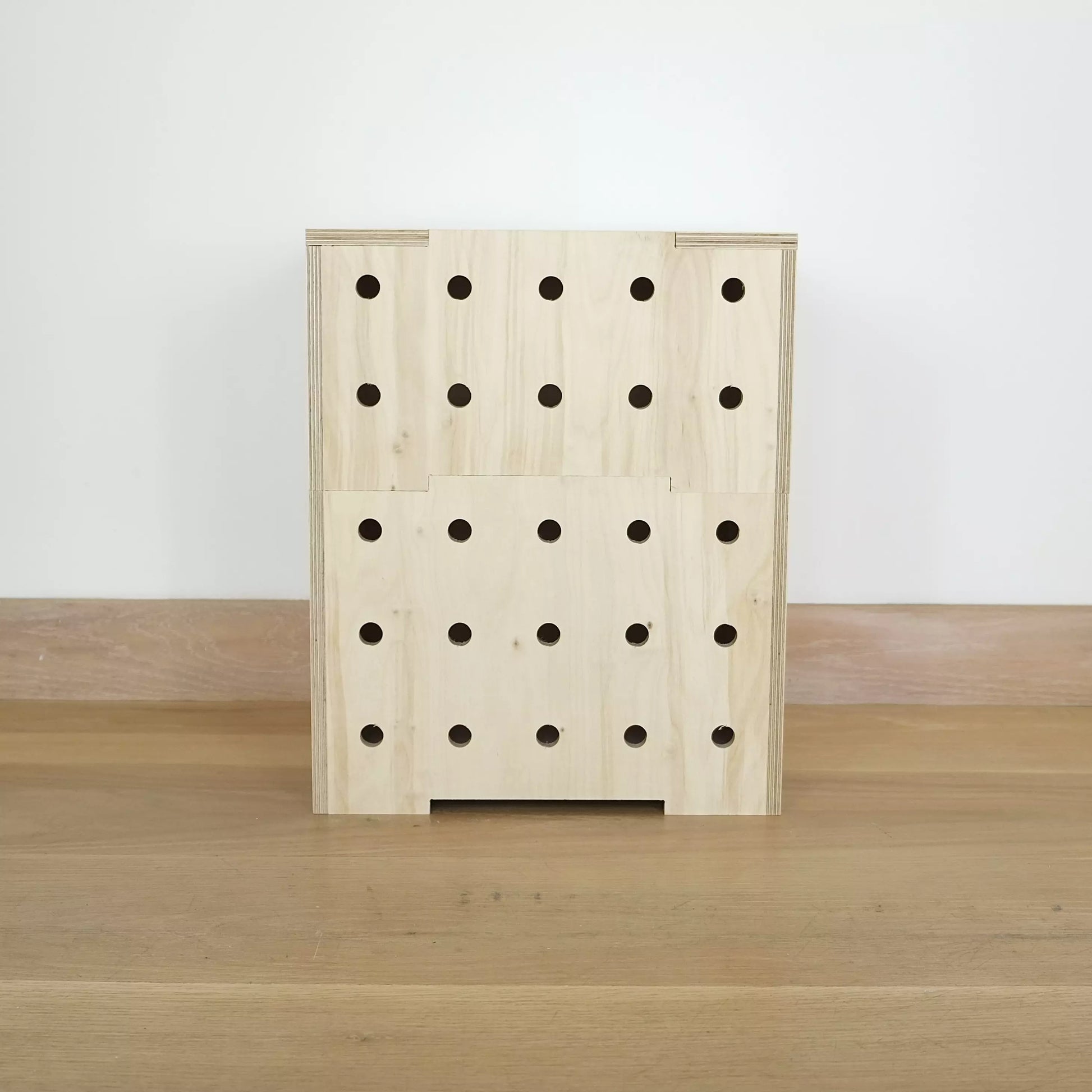 Stack of two simple pale wooden square shaped storage crates facing front on, with five rows of holes cut in the front facet and a wooden lid, sitting on a wooden floor.